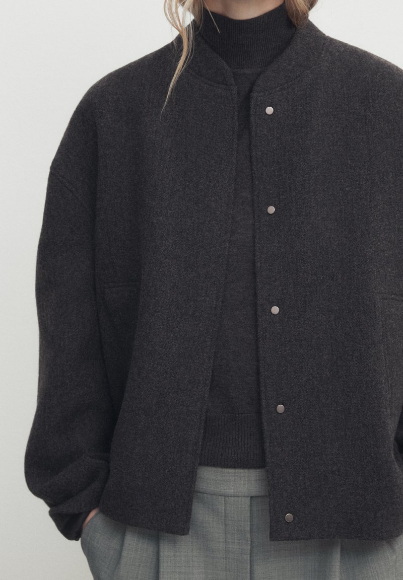 Veste en mélange de laine gris foncé avec un col rond, des fermetures à pression et une coupe décontractée. Portée sur un pull assorti de couleur sombre, associée à un pantalon gris clair.