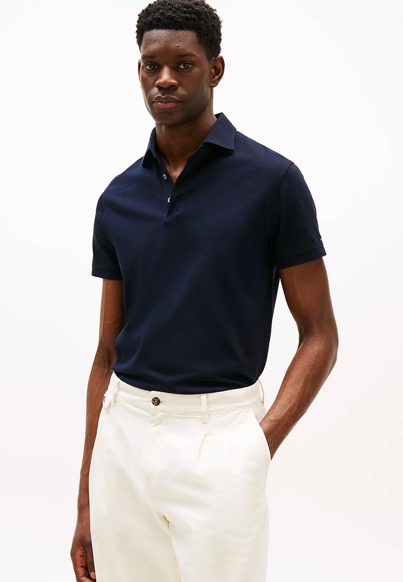 Polo shirt blu navy con colletto classico e tre bottoni, realizzato in tessuto liscio. Indossato con pantaloni di colore chiaro.