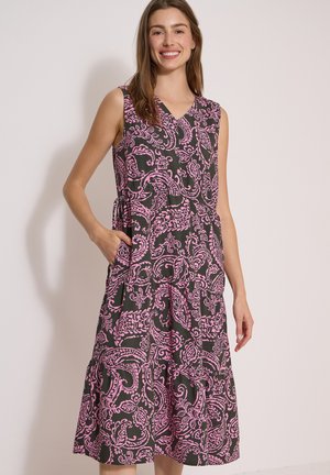 Femme souriante portant une robe noire sans manches avec de grands motifs cachemire roses, une main dans la poche, debout devant un fond clair uni.