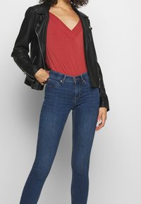 Giacca di pelle nera sopra un top rosso a V; jeans in denim blu attillati; texture morbide, ensemble casual che mette in risalto un vivace contrasto di colori.