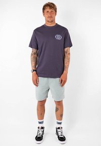 T-shirt à manches courtes de couleur violette foncée avec un motif floral sur la poitrine, associé à un short vert clair et des chaussures de skate noires.