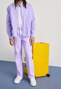 Lila košile a kalhoty z bavlněné látky, doplněné bílými teniskami. V pozadí je žlutý pojízdný vozík.