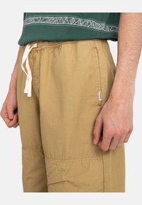 Homme portant un pantalon beige à cordon de serrage et une chemise verte avec des bordures à motifs, les mains relaxées sur les côtés, visible de la taille à mi-cuisse.
