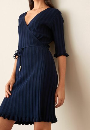 Femme portant une robe portefeuille en maille côtelée bleu marine avec des manches trois-quarts, une ceinture à nouer et un ourlet volanté, debout contre un mur clair.