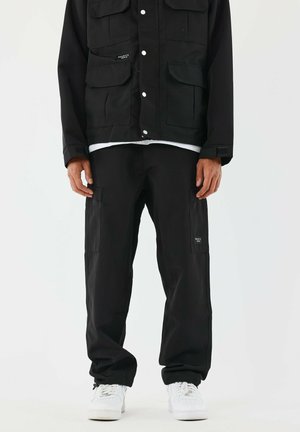 Pantalones cargo negros con múltiples bolsillos, combinados con una chaqueta negra a juego con botones a presión y una tela texturizada. Zapatillas blancas.