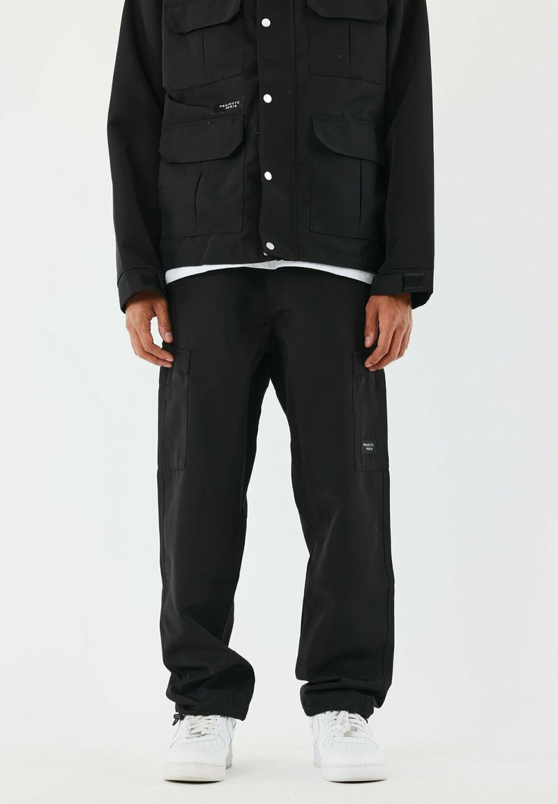 Pantaloni cargo neri con molte tasche, abbinati a una giacca nera coordinata con bottoni a pressione e un tessuto testurizzato. Sneakers bianche.