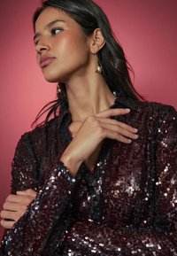 Pitsiliste litritega kaunistatud burgundiapunane pluus kraega, millel on tekstureeritud pind ja pikad varrukad, esitletud roosa tausta ees.