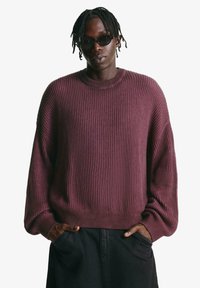 Maroon ribbed knit sweater with a round neck and loose-fitting sleeves. Paired with black pants, hands rest in pockets. Textured fabric.