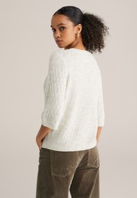 Pulover alb, texturat, cu model în formă de diamant, cu mâneci trei sferturi. Asortat cu pantaloni din catifea de culoare verde măsliniu, vedere din spate.