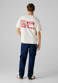 Hvid bomulds t-shirt med fed rød tekst: "RED BULL RACING WORLDWIDE." Sammen med blå denim jeans, afslappet pasform, og hvide sneakers.