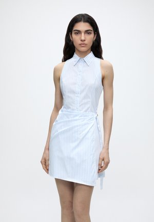 Mladá žena s dlhými tmavými vlasmi, ktorá má na sebe svetlomodré, bezrukávové šaty s vertikálnymi pruhmi, stojaca pred jednoduchým bielym pozadím.