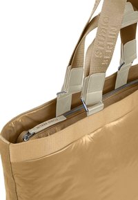 Beige tote bag med en glatt, blank overflate, med doble skulderremmer og glidelås på toppen. Inkluderer en etikett med "STUDIO BERLIN" preget.
