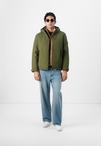 Grüne Kapuzenjacke mit einem Reißverschluss vorne, getragen über einem braunen Shirt. Kombiniert mit hellblauen weit geschnittenen Jeans und weißen Sneakers. Runde Sonnenbrille.