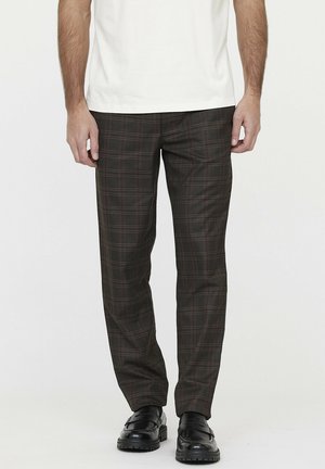 Mężczyzna w ciemnobrązowych spodniach w kratę, białej koszuli i czarnych mokasynach, stojący na tle jasnego, jednolitego tła.