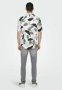 Camisa blanca de manga corta con un patrón de hojas tropicales en verde y negro, combinada con unos vaqueros tapered de color gris claro y zapatillas deportivas blancas.