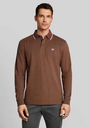 Man wearing a brown long-sleeve polo shirt with a striped collar and dark gray pants standing against a plain background.