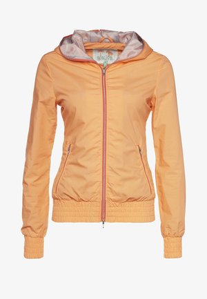 Chaqueta de color naranja claro con forro gris en contraste, jeans oscuros de corte ajustado y zapatillas deportivas negras con acentos de color, llevando una bolsa oscura.