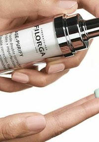 A silver and white bottle labeled "FILORGA AGE-PURIFY" is held over a hand, dispensing a small amount of light green fluid.