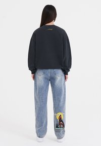 Zwarte, oversized sweatshirt met een ronde hals, voorzien van een kleine gouden tekstdetail op de achterkant. Lichtblauwe, relaxte spijkerbroek met een wolfgrafiek.
