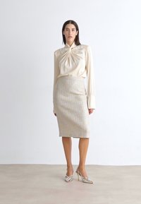 Lys beige bluse med knyttet frontdetalje, parret med en tætsiddende, mønstret blyantskirt i lyse pastelfarver, og sølvfarvede højhælede sko.