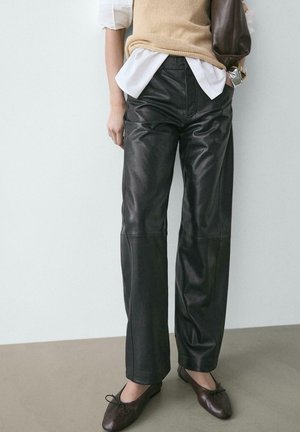 Donna che indossa pantaloni in pelle neri, ballerine marroni, un gilet beige lavorato a maglia sopra una camicia bianca, e porta una borsa a tracolla marrone scuro.