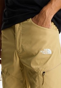 Mano nella tasca dei pantaloncini beige The North Face con dettagli a cerniera, indossati con una maglietta nera.