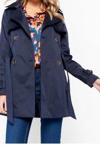 Mörkblå trenchcoat med dubbelknäppning, guldfärgade knappar och klassisk krage, bärs över en färgglad blommig blus.