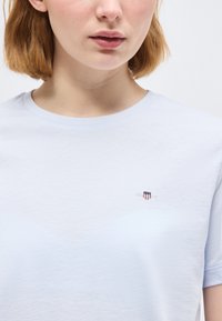 Άτομο με ανοιχτόχρωμο δέρμα και κοντά κόκκινα μαλλιά, που φοράει μια ανοιχτά γαλάζια μπλούζα με στρογγυλή λαιμόκοψη και μικρό λογότυπο ασπίδας στο αριστερό στήθος.