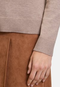 Maglione beige lavorato a maglia con orlo e polsini a coste, indossato su una gonna in suede marrone. Ingrandimento di una mano appoggiata dettagliatamente sul tessuto.
