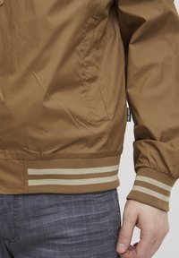 Smeđa najlonska bomber jakna s rebrastim ovratnikom i manžetama, s naglašenim prugama. Uključuje bočni džep i vidljive detalje šivanja.