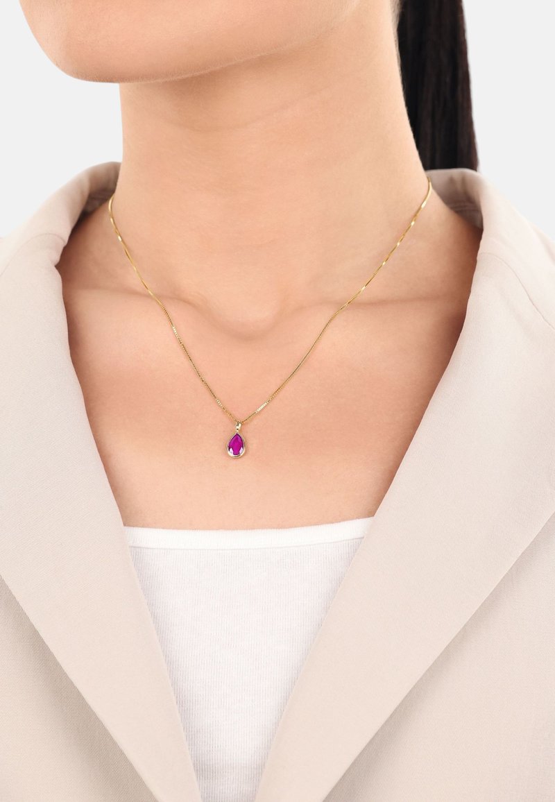 Goldene Anhängerkette mit einem tropfenförmigen lila Edelstein, der an einer dünnen Kette hängt, getragen über einem weißen Oberteil und unter einem beigen Blazer.