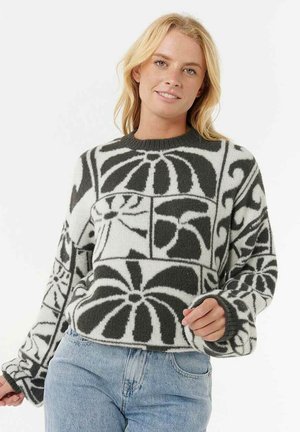 Donna bionda che indossa un maglione a righe bianche e nere con design floreali astratti, abbinato a jeans azzurri, mentre sorride verso la fotocamera.