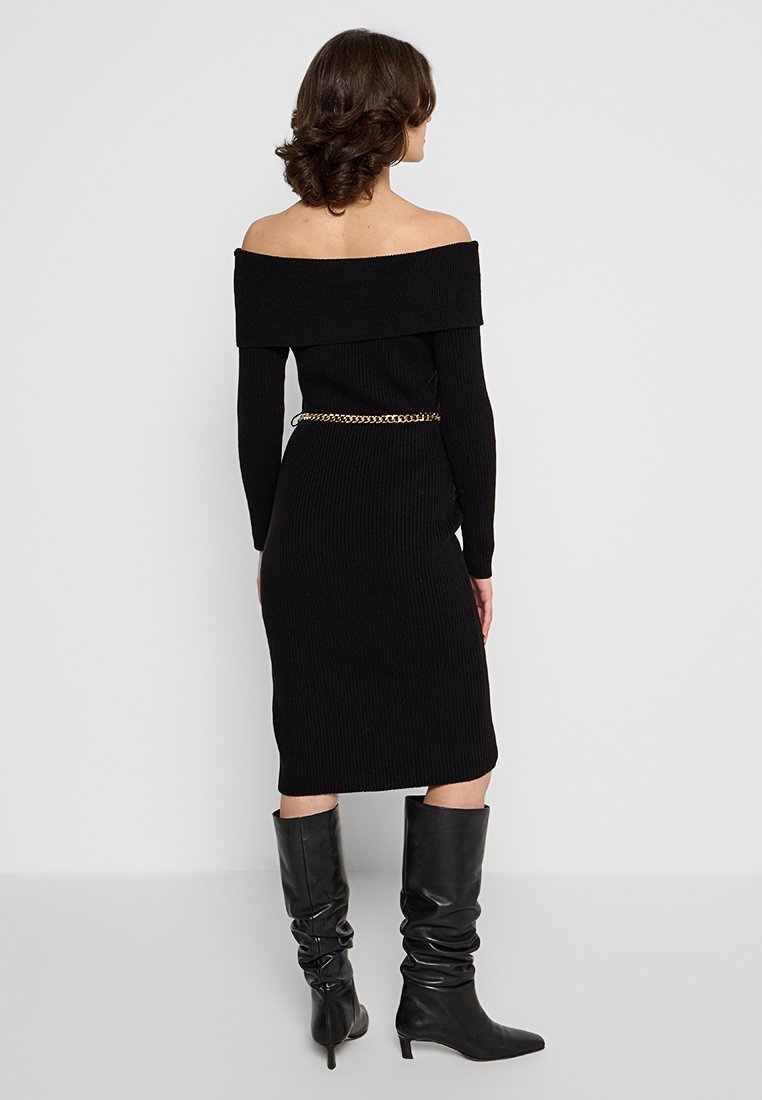 Abito nero a coste con design off-shoulder, forma aderente e lunghezza al ginocchio. Accentuato da una cintura in catena dorata e abbinato a stivali neri al ginocchio.
