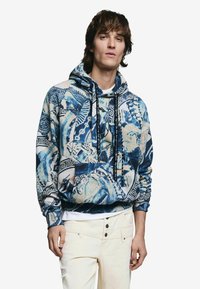 Modrá a smotanová mikina s kapucňou a zložitými vzormi. Bavlnený materiál, voľný strih, s predným vreckom a čiernymi šnúrkami na stiahnutie.