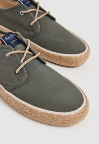 Zapatos de lona verde oscuro con detalles de cuerda, punta redonda y suela tejida beige. Presentan cordones beige y una etiqueta con el logo en azul marino en la lengüeta.