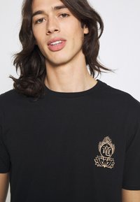 T-shirt preta de algodão com um pequeno desenho dourado contendo um motivo intricado e texto na área do peito esquerdo. Decote em crew clássico.
