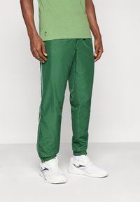 Pantalones deportivos verde ligero con rayas blancas en los costados, puños elásticos y textura suave, combinados con zapatillas blancas.