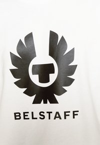 Camiseta blanca con un gran logotipo negro con alas sobre un emblema circular y la palabra "BELSTAFF" debajo en letras negritas negras.