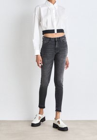 Persona vistiendo una camisa blanca corta de botones, jeans oscuros de cintura alta ajustados con dobladillos deshilachados, y zapatos blancos gruesos con suelas negras.