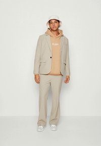Chaqueta de traje beige a cuadros con pantalones a juego, superpuesta sobre una sudadera con capucha de color marrón claro. Llevada con un sombrero tipo bucket blanco y zapatillas grises.
