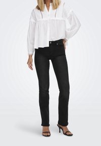 Blouse blanche avec rayures verticales texturées et boutons sur le devant, associée à un jean foncé. La tenue comprend des talons noirs à lanières.