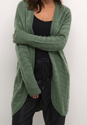 Femme portant un cardigan en maille vert sur un haut noir et un pantalon noir à cordon, debout les bras croisés devant un fond uni.