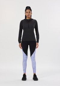 Schwarzes langärmliges Oberteil mit halbem Reißverschluss und eng anliegender schwarzer Leggings mit lila Marmor-Muster an den Seiten. Das Model trägt schwarze Sportschuhe.