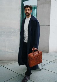 Navy overcoat, white sweater, black pants, brown leather briefcase, dark brown shoes, set against a light concrete background.
