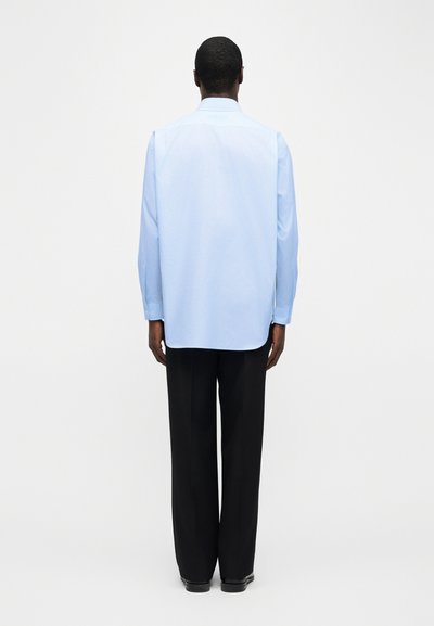 Άνδρας που στέκεται με την πλάτη, φοράει ανοιχτό μπλε πουκάμισο με μακριά μανίκια και μαύρο παντελόνι με φαρδιά μπατζάκια σε απλό λευκό φόντο.