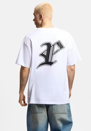 Osoba z krótkimi blond włosami, ubrana w białą koszulkę z dużą czarną stylizowaną literą „P” na plecach oraz niebieskie dżinsy.