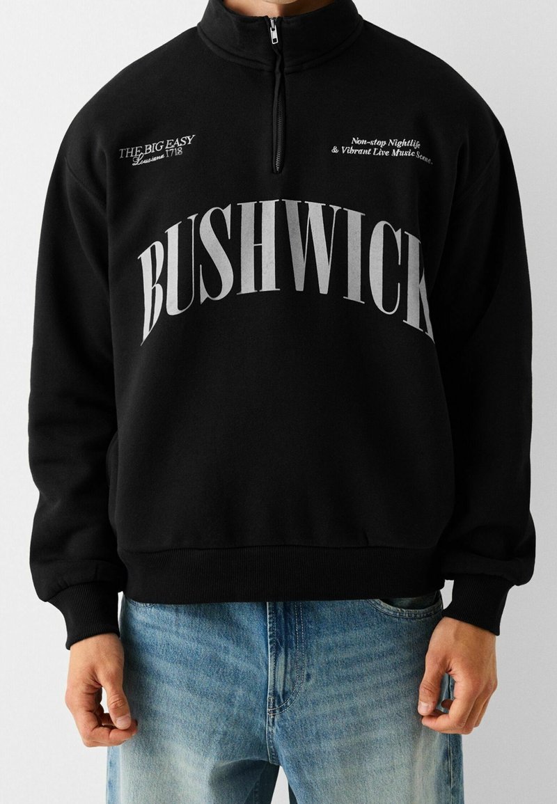 Svart zip-upp sweatshirt med stora vita bokstäver "BUSHWICK", ytterligare text i mindre teckensnitt och ribbade muddar och nederkant.