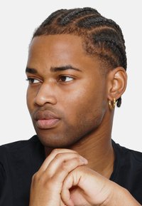 Un jeune homme aux cheveux tressés et foncés, portant une chemise noire et une boucle d'oreille en or, a la peau lisse et une expression neutre.