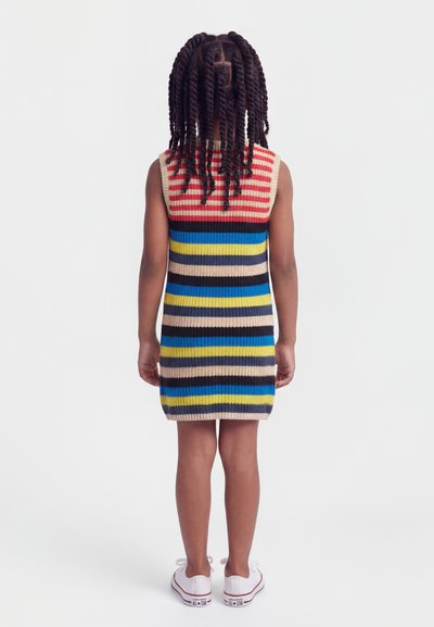 Παιδί με κοτσίδες που φοράει ένα αμάνικο, πολύχρωμο ριγέ πλεκτό φόρεμα και λευκά αθλητικά παπούτσια, στέκεται με την πλάτη γυρισμένη σε λευκό φόντο.