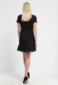 Abito nero corto con maniche corte, caratterizzato da un bordo in pizzo sulle maniche e sull'orlo. Indossato con tacchi alti neri, che mettono in risalto una silhouette svasata.
